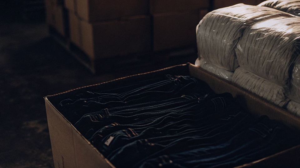 A large, compressed bale of raw cotton sitting next to an open cardboard box neatly filled with folded jeans.