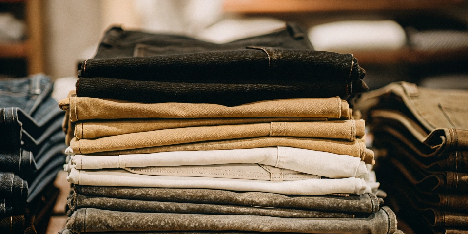 A stack of jeans neatly folded to show off a variety of modern colors like black, grey, olive green, and beige.