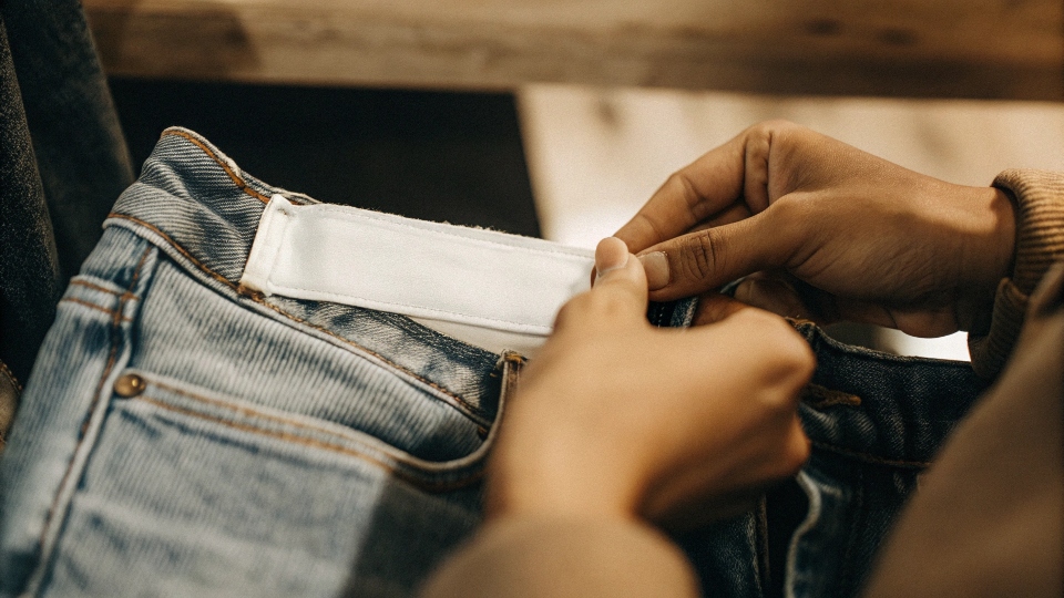A close-up photo of a person's hands checking the white care label inside the waistband of a pair of blue jeans.
