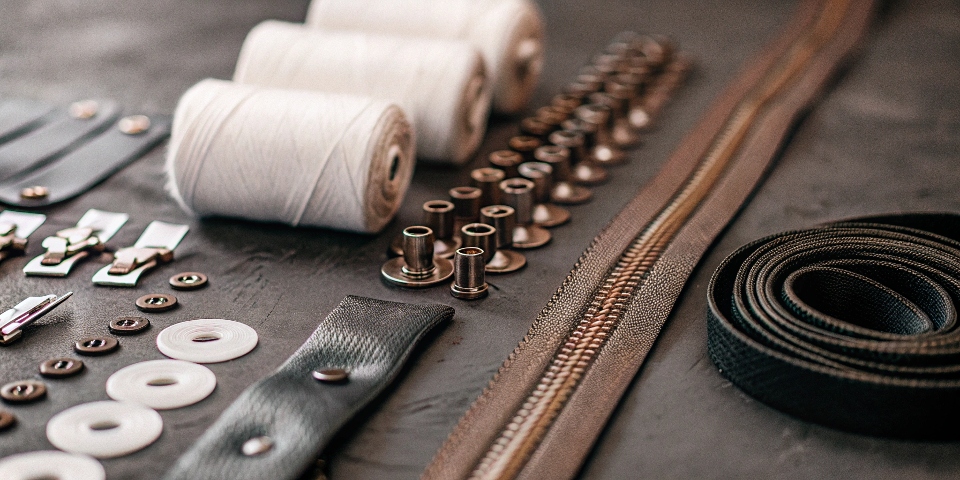 An artfully arranged flat lay of raw materials: a roll of denim, buttons, rivets, a zipper, and spools of thread.