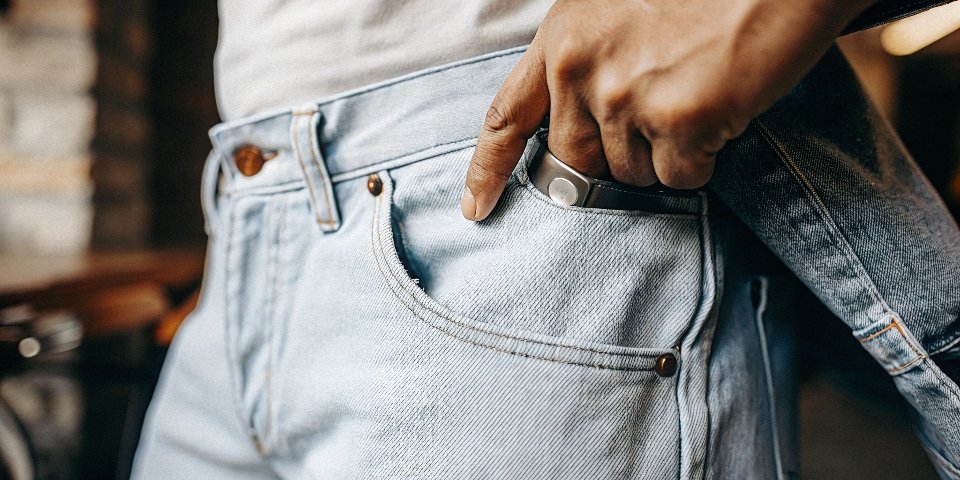 A close-up shot of a person's hand pointing specifically to the small fifth coin pocket and the copper rivet on a pair of blue jeans.