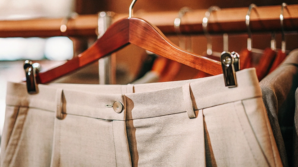 A close-up shot of elegant grey wool trousers hanging on a wooden hanger, highlighting the sharp center crease and fine fabric texture.