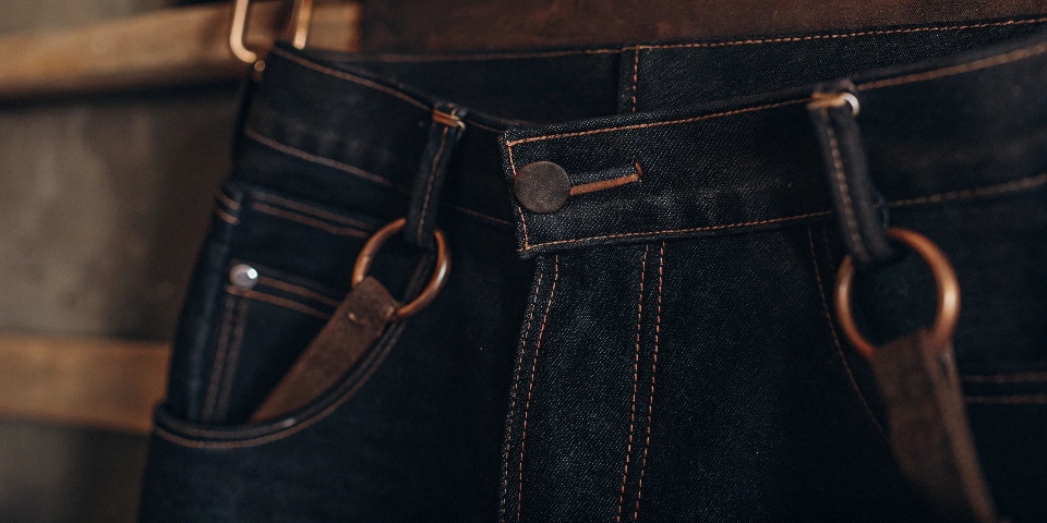 A detailed shot highlighting premium jeans features: a thick leather patch, chain-stitched hem, and custom copper hardware.