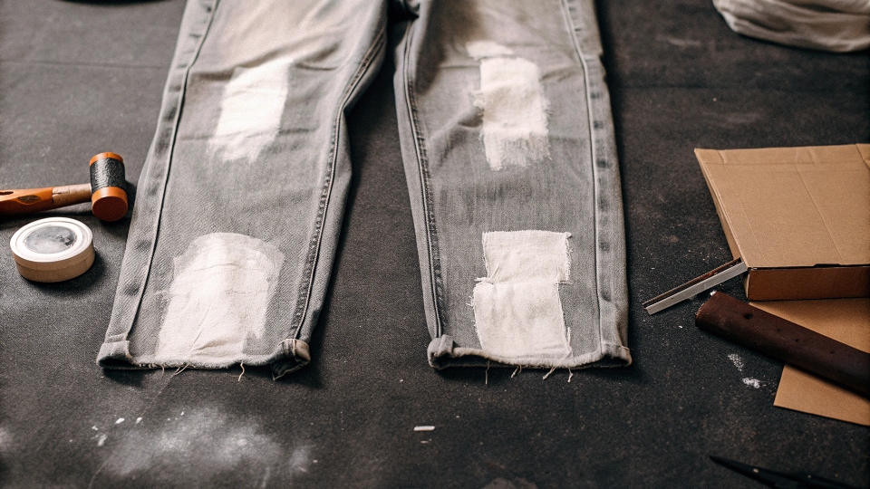 A DIY setup showing jeans with chalk marks, a razor, sandpaper, and a piece of cardboard.
