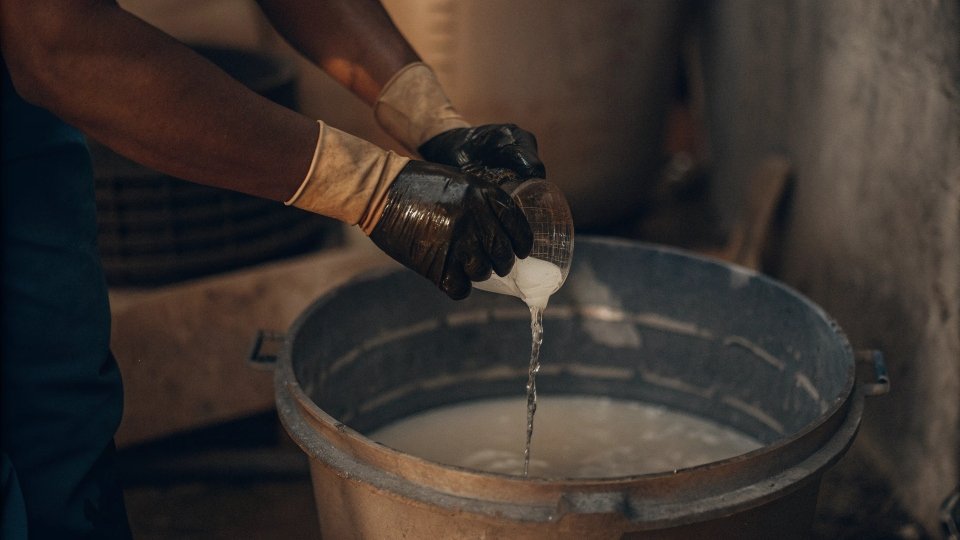 A person wearing gloves carefully pouring bleach from a measuring cup into a large bucket of water.
