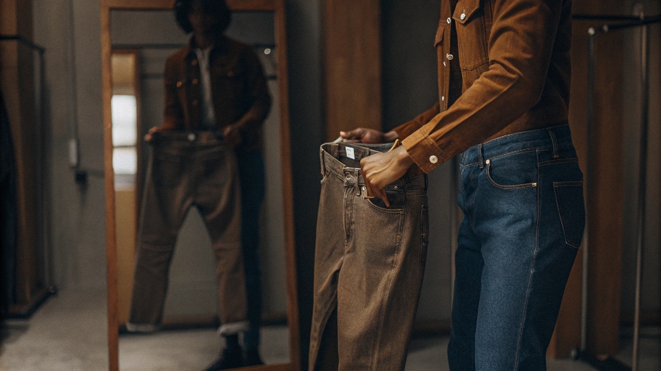 A person in a fitting room holding two pairs of jeans, looking thoughtfully at the size tags.