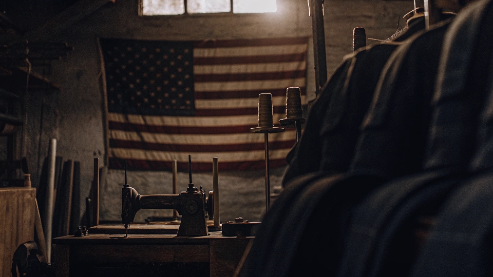 An American flag hanging in the background of a small-scale denim workshop, with bolts of selvedge denim and sewing machines visible.