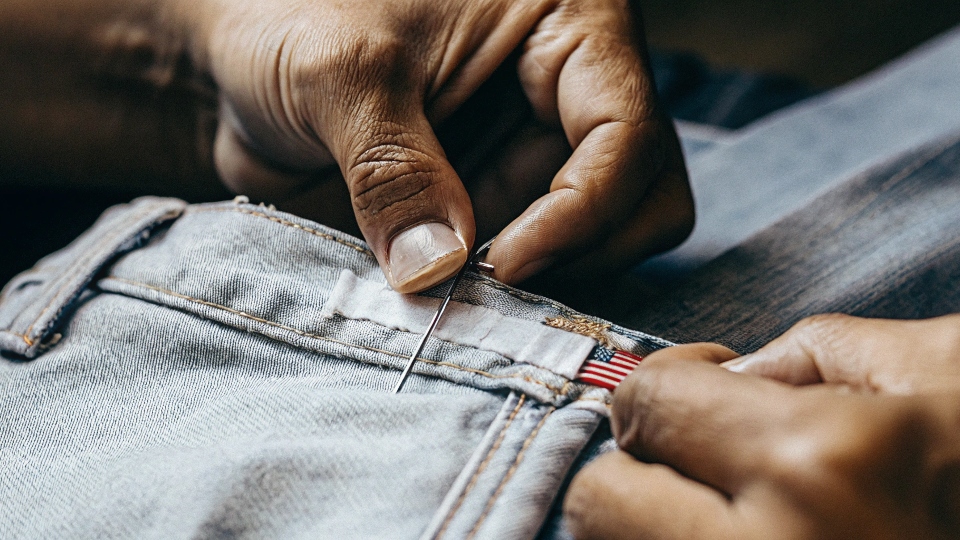 A close-up of hands sewing a "Made in USA" label onto a pair of jeans.