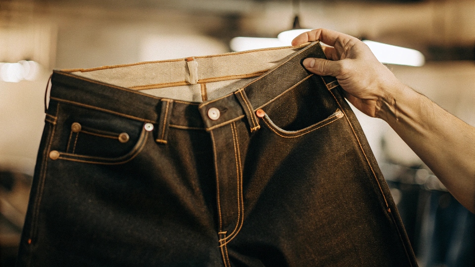 A hand holding up a pair of high-quality raw denim jeans, with light shining through to show the dense texture.