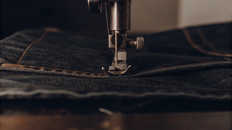 A sewing machine needle stitching a new seam along the inseam of an inside-out pair of jeans.