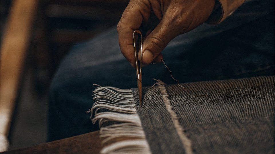 A close-up shot of a jean hem, showing the white horizontal threads being pulled out by tweezers to create fringe.