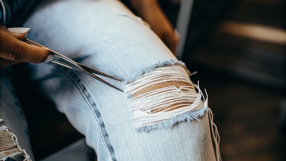 A close-up of tweezers pulling blue threads from a jean's knee to create a distressed look.