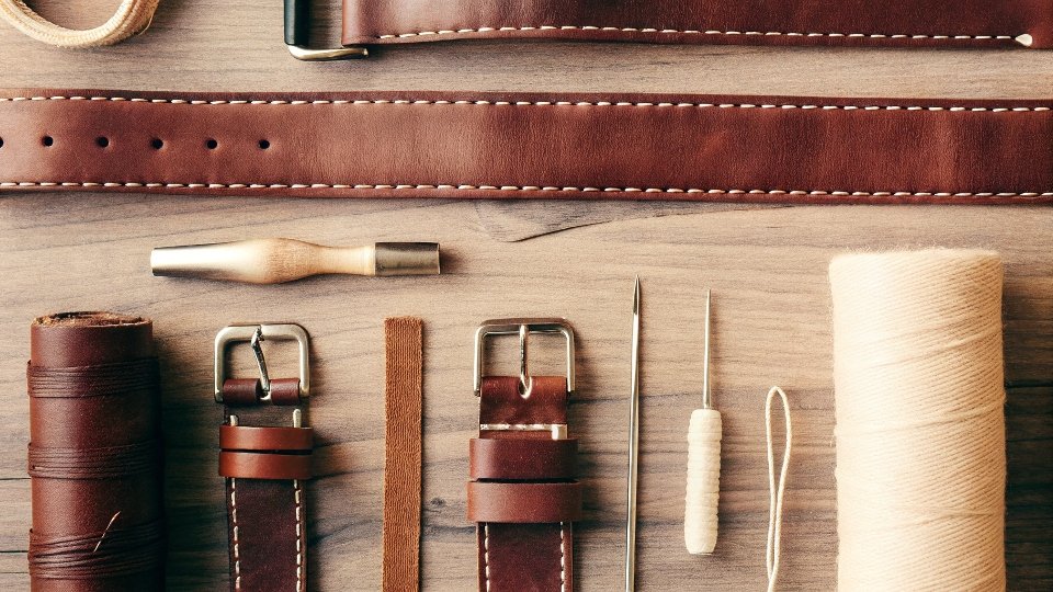 An overhead shot of various tools for altering a jean waistband laid out on a denim background: a needle, thread, elastic, a shoelace, and waist adjustment clips.