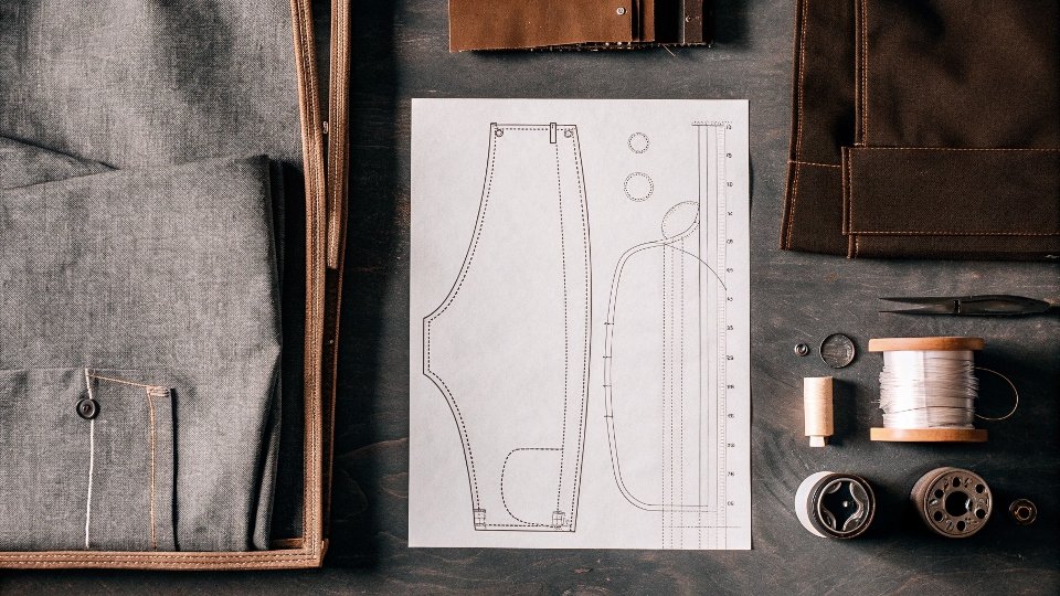 A flat lay of essential jean-making supplies: denim fabric, a pattern, thread, needles, and hardware.