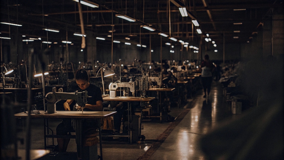 A bustling, bright, and modern sewing floor in a Los Angeles garment factory, with workers operating industrial sewing machines.