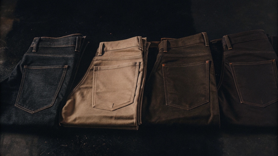 An assortment of five-pocket style pants in different materials: one in denim, one in khaki-colored canvas, and one in brown corduroy.