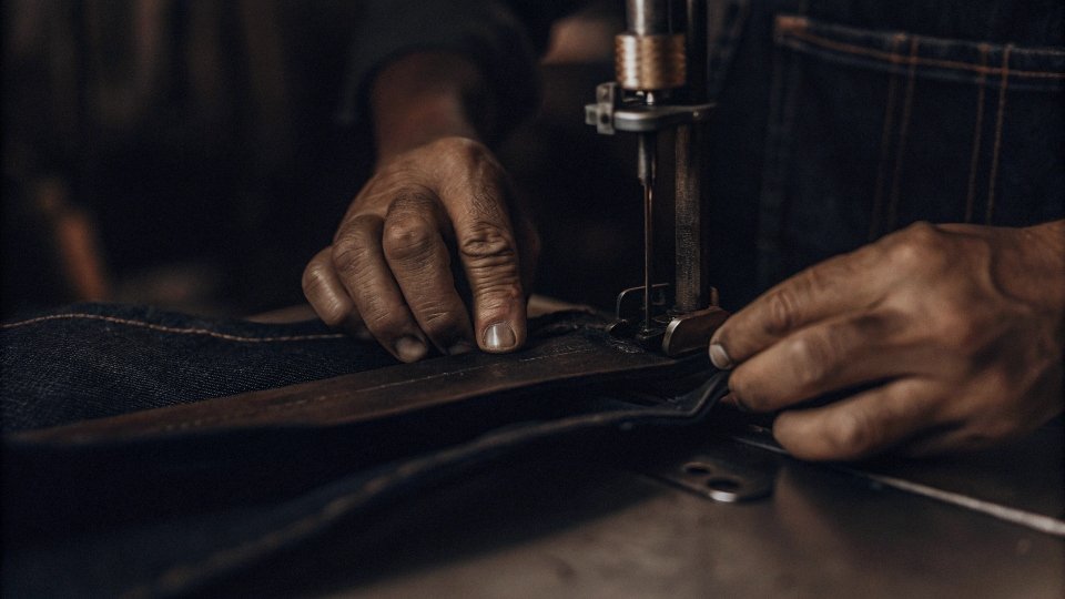 A tailor's hands shown working on the center back seam of a pair of jeans, with the belt loop removed.