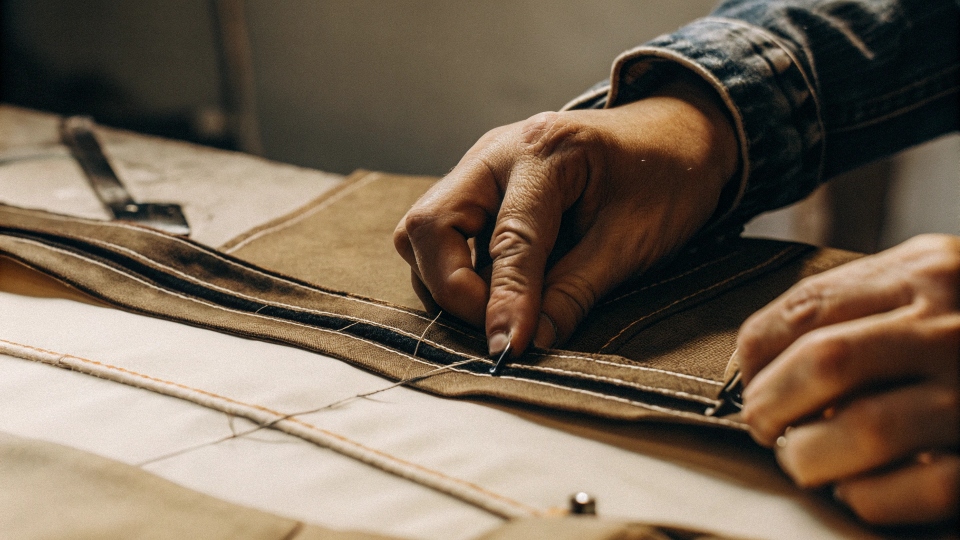 A tailor's hands carefully unpicking the stitches of a jean's waistband, showing the start of the deconstruction process.