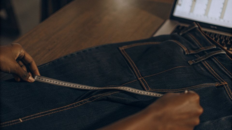 A person using a measuring tape on a pair of jeans, with a size chart visible on a laptop screen nearby