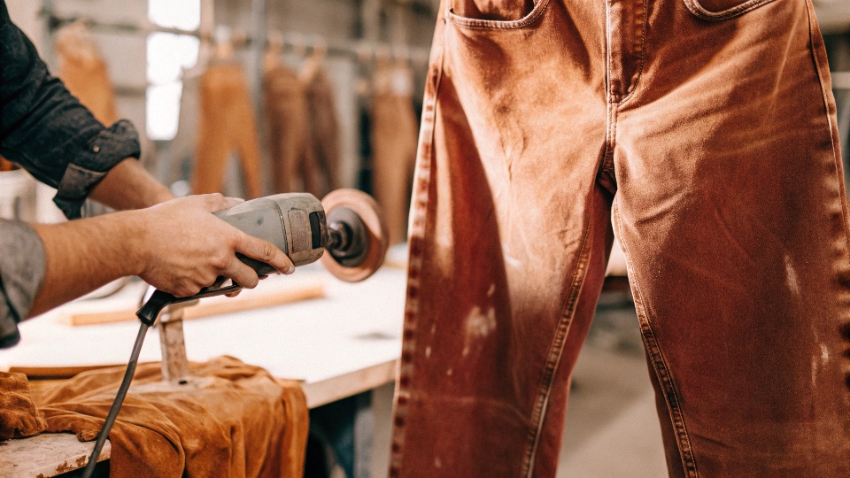 The Process of Creating Distressed Jeans A denim artisan in a factory using a handheld sander to create fades on the thigh of a jean