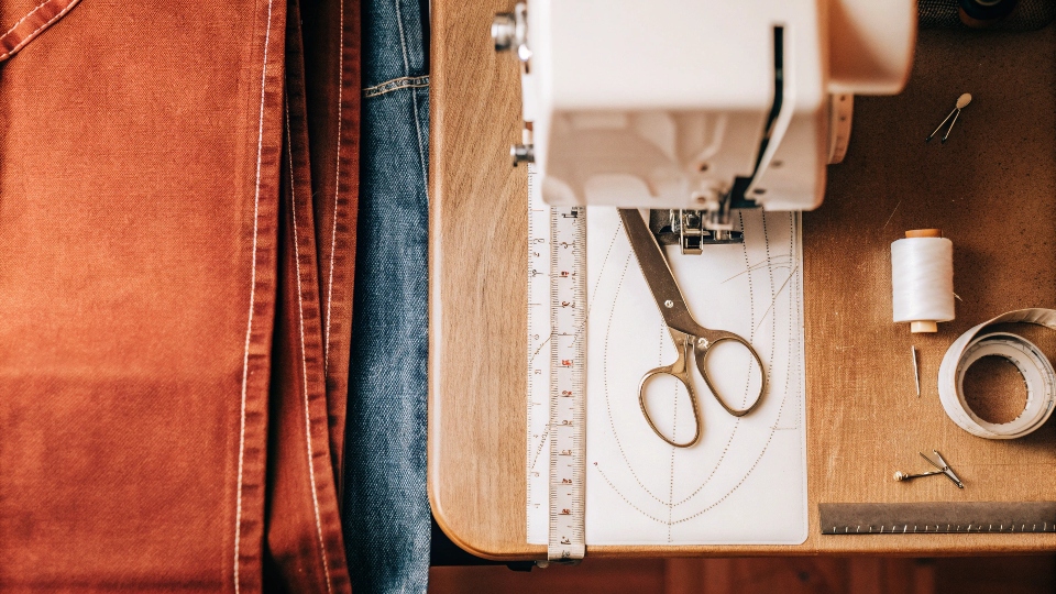 A top-down view of a sewing workspace with denim fabric, a pattern, scissors, and a [sewing machine](https://diznewjeans.com/how-to-make-your-own-jeans/)[^3]