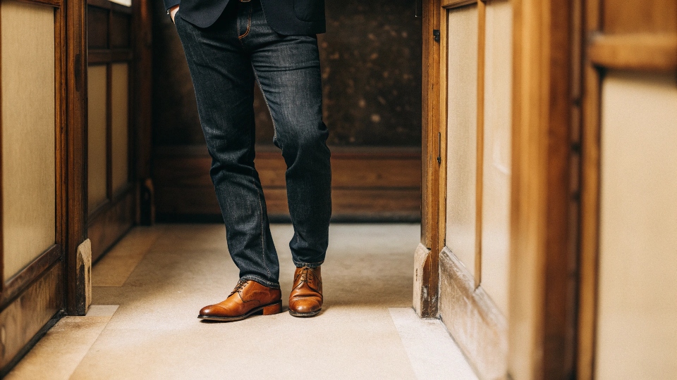 A well-dressed person wearing dark, tailored jeans with a sharp navy blazer and brown leather brogues