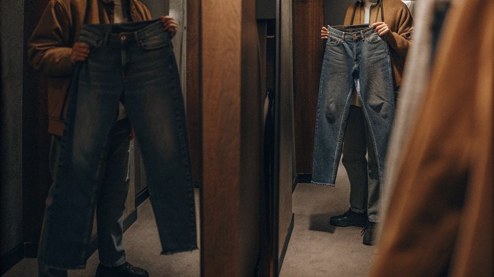 A person in a fitting room looking unsure while deciding between two sizes of jeans