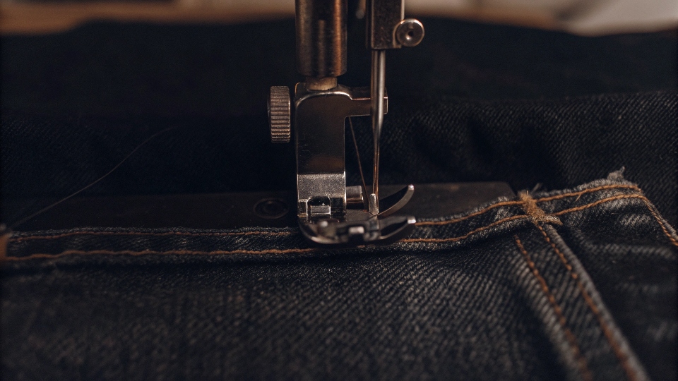 A close-up shot of a sewing machine needle stitching a dart on the waistband of jean shorts