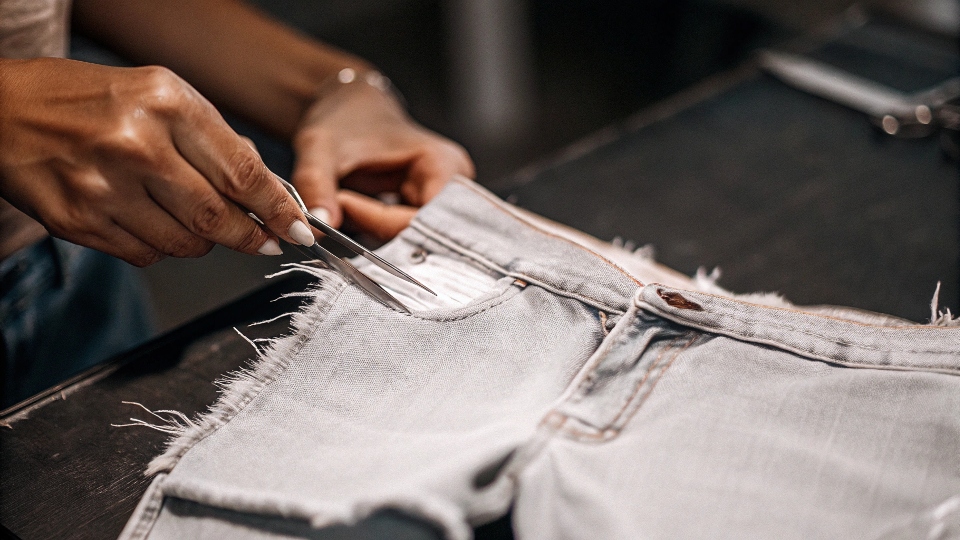 A person using tweezers to pull at the horizontal threads on the edge of cut-off jean shorts