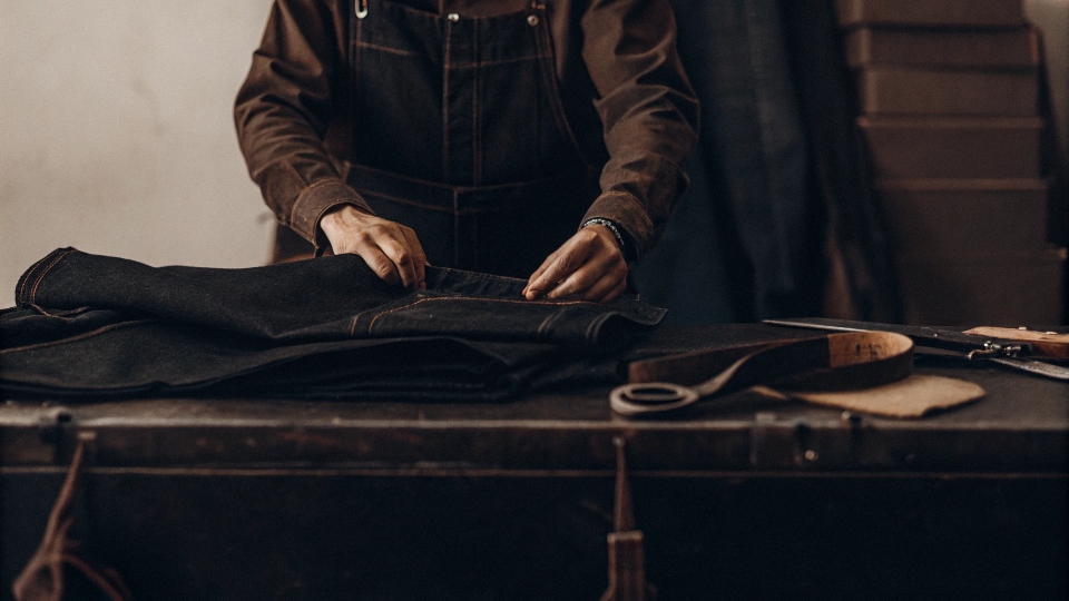 A designer carefully examining a pair of jeans on a workbench with fabric swatches nearby