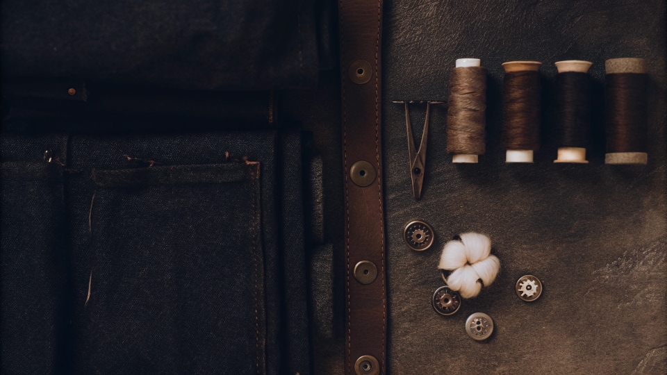 A flat lay of raw materials: a bolt of denim, cotton bolls, spools of thread, rivets, and buttons