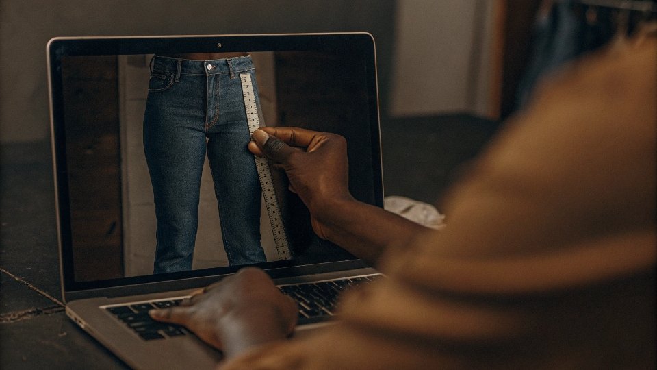 A person comparing their handwritten measurements on a notepad to an online retailer's detailed jean size chart on a laptop screen