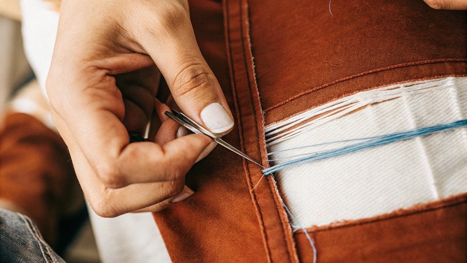 A close-up of a person using tweezers to pull blue threads from a cut in a pair of jeans