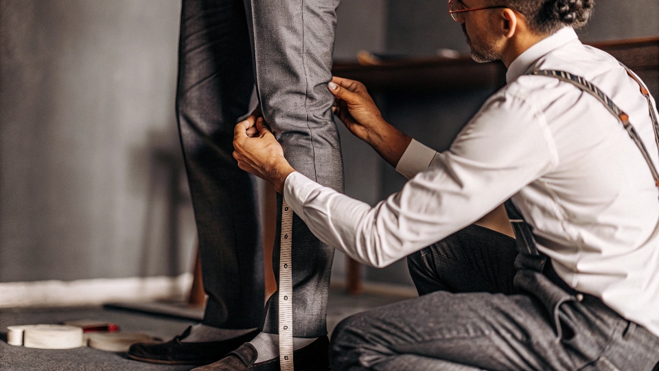 A tailor on one knee, carefully pinning the inseam of a customer's jeans to mark for a skinny taper