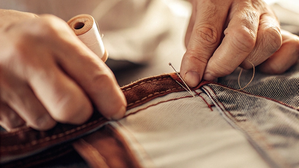 A close-up of a tailor's hands working on the center back seam of a jean's waistband