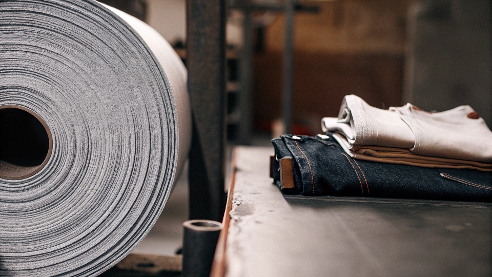 An image showing a large roll of denim fabric on one side and a finished pair of folded jeans on the other