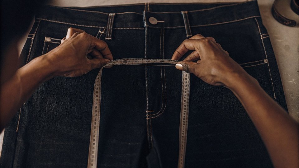 A [tailor](https://diznewjeans.com/can-a-tailor-really-make-a-jeans-waist-smaller/)[^2]'s hands measuring the rise of a pair of jeans with a tape measure