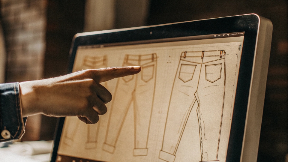 Online Digital Proofing of a Jeans Tech Pack A designer's hand pointing to a detail on a computer screen showing a digital 'tech pack' of a pair of jeans.