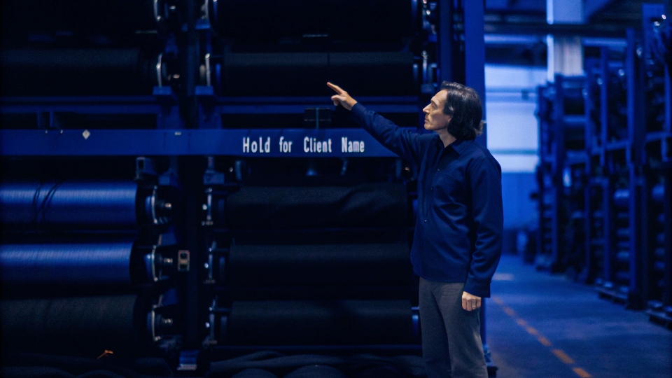 A factory manager pointing to a roll of fabric marked 'Hold for [Client Name]' in a warehouse.