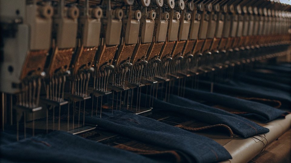 Multi-Head Computerized Embroidery Machines A row of modern, multi-head Tajima computerized embroidery machines stitching the same logo on multiple jeans at once.