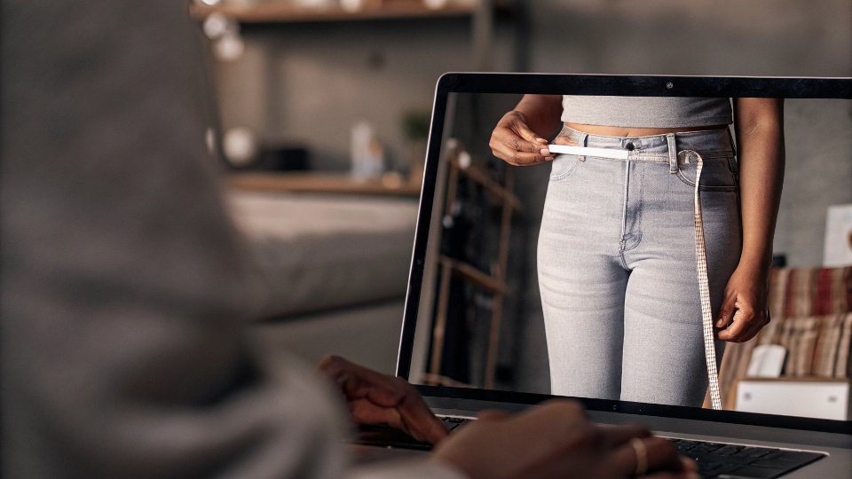 Capturing Detailed Measurements on Video A close-up shot on a video call showing a technician's hands measuring the waist of a pair of jeans on a model, with the tape measure clearly visible.