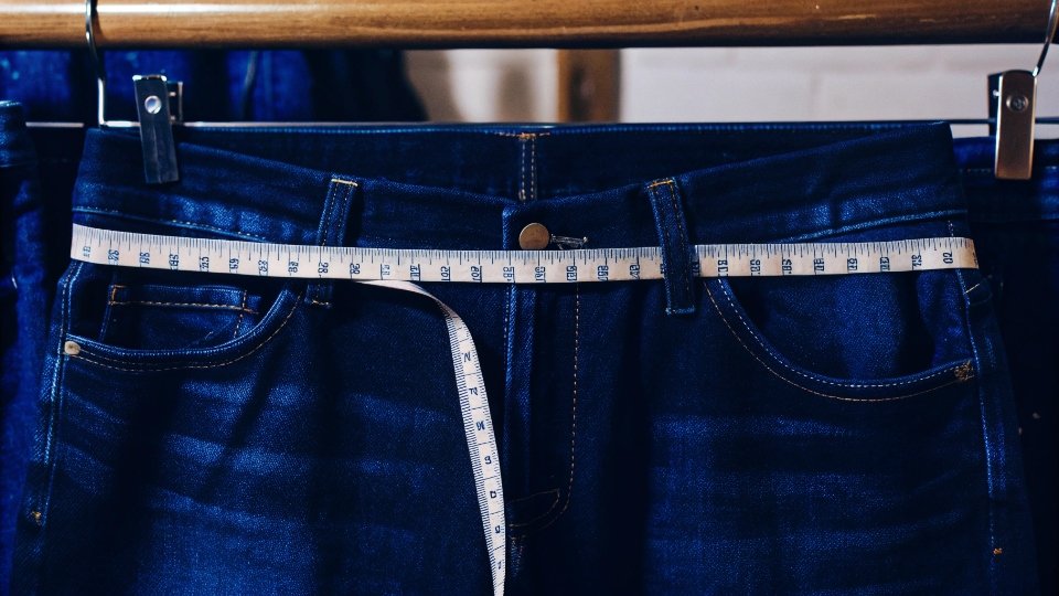 Measuring Jeans Shrinkage in Sizes A tailor's measuring tape showing a one-inch reduction on the waistband of a pair of jeans.