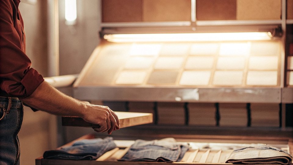 Comparing RFH Sample Color to Bulk Production Under a Lightbox A quality control inspector holding an RFH jean sample next to a jean from the bulk production line, using a lightbox to compare colors.