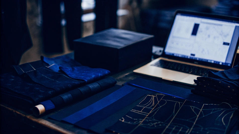 The Elements of a Private Label Clothing Brand A designer's desk with fabric swatches, sketches, a laptop showing an e-commerce site, and a branded shipping box.