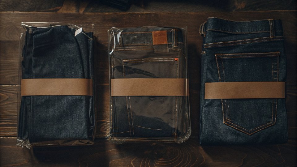 Bundled Jeans Multi-Pack and Set Packaging A 3-pack of jeans neatly held together with a branded paper belly band, next to a denim jacket and jeans set folded together in one large polybag.
