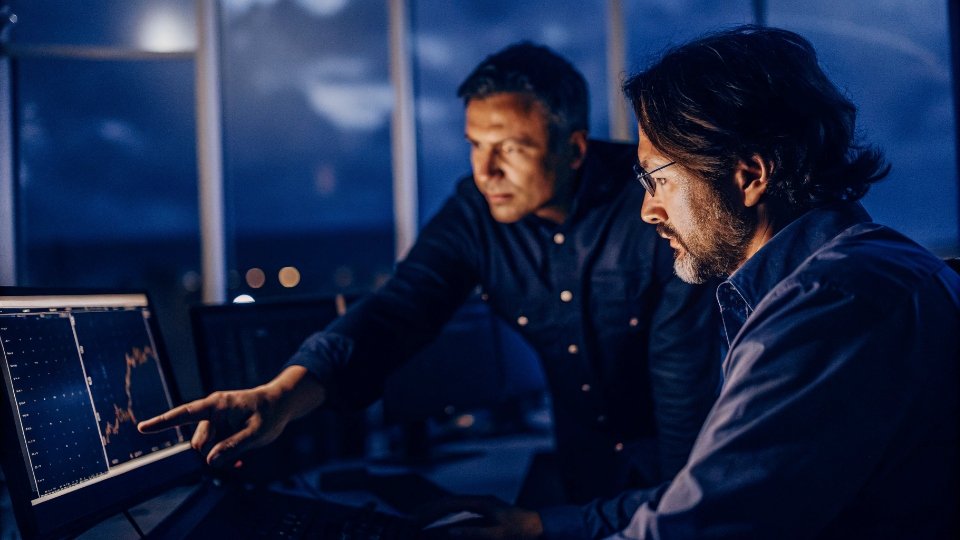 Mid-Season Forecast Adjustments A designer on a laptop sharing a sales graph with the factory manager, who adjusts a production schedule.