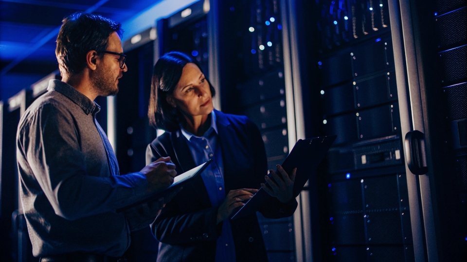 Auditing IT Security Practices An IT professional with a clipboard reviewing security protocols with a factory manager in front of a server.