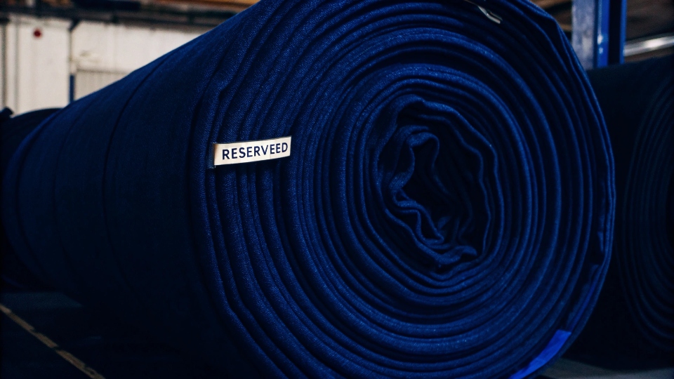 Locking Fabric Costs with Advance Purchase A large roll of denim fabric in a warehouse with a 'Reserved' sign on it.