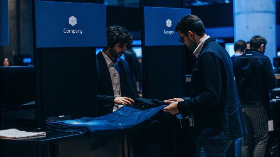 Co-Branded Trade Fair Booth Two company logos on the wall of a trade show booth, with a designer showing jeans to a potential buyer.