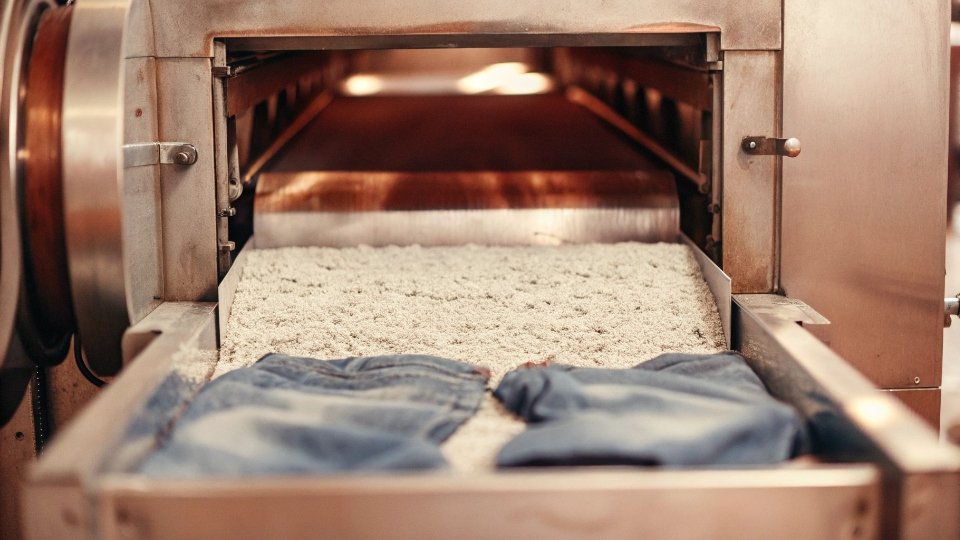 The Industrial Process of Acid Washing Jeans An industrial laundry machine with its door open, showing a mix of jeans and pumice stones inside.